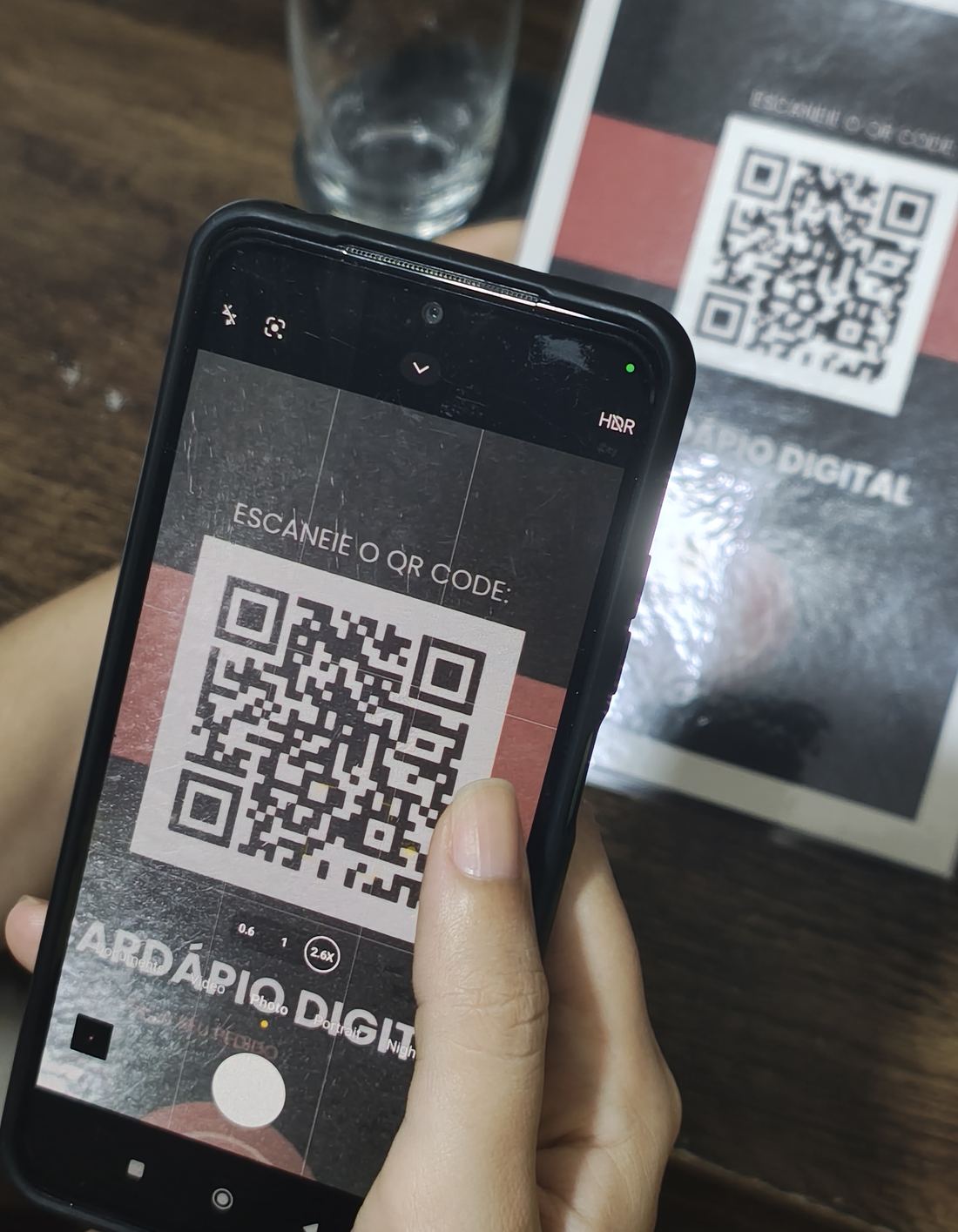 Mão apontando a câmera do celular para um cartão impresso na mesa com o QR Code do cardápio digital